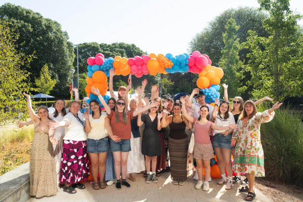 Students looking to camera smiling and waving with balloon arch in the background