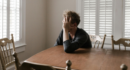 Man sat at a wooden table looking towards the nearby window with his head in his hand