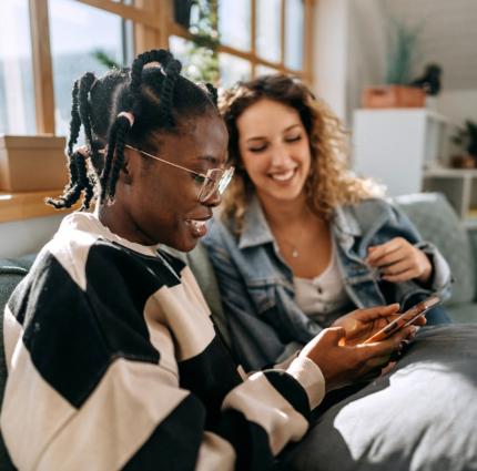 Two young women sat together on a sofa laughing and smiling whilst looking at something on their phone together