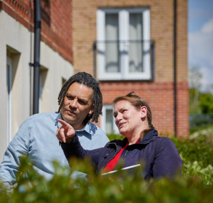 Man with dreads is looking off towards a building whilst his female colleague holds a clipboard and is pointing at something of interest