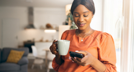 Young black lady wearing an orange top is standing looking at her phone whilst having a hot drink in a white mug