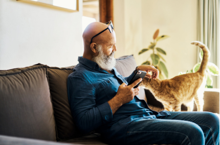 Bald man with a white beard and clack reading glasses resting on top of his head is sitting on a sofa holding his phone and stroking a small ginger cat next to him