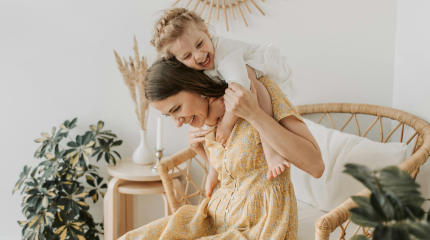 Young mum sitting on a wicker chair with her young daughter climbing into her shoulder giggling