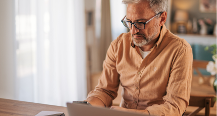 A man wearing glasses and a shirt is sitting at a table with his laptop open and looking at his phone