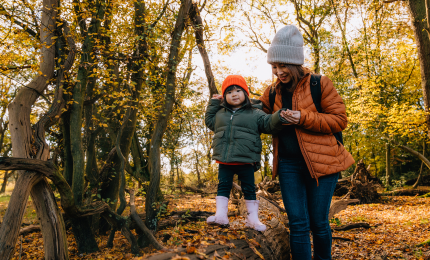 Young girl is looking straight ahead as she balances on a tree trunk that her mum is helping her across. They are in a forest with autumnal leaves all around them.