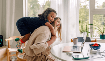 Young mum sat at her laptop at a table with her daughter climbing up and hugging her around her shoulders