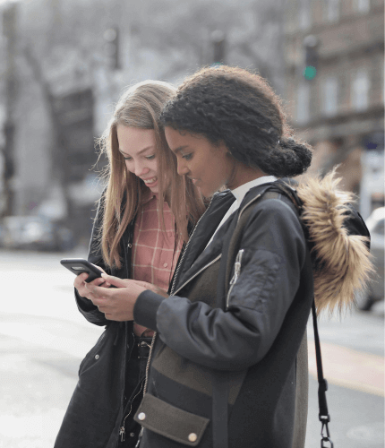 Image shows two young individuals smiling and looking at a phone together outside on a pavement