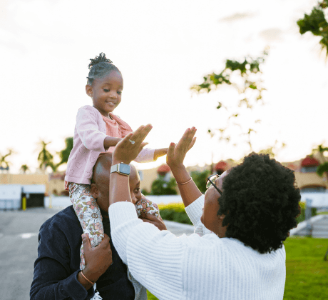 Child sitting on their parents shoulders whilst giving a double high five to their other parent