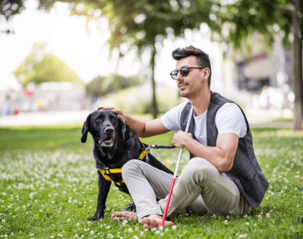 Blind customer sits on a grassy field with their guide dog sitting beside them