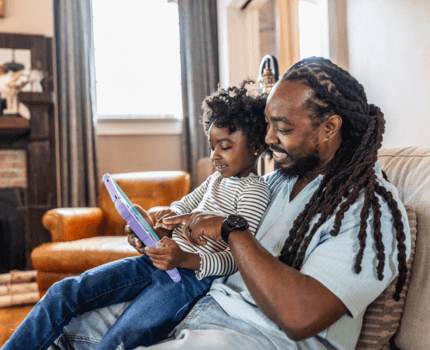 Young dad sits on a sofa whilst his daughter sits on his lap and they both use her tablet