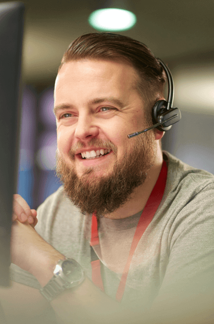 Image shows a close up of someone with short hair, smiling and speaking into a headset in front of a computer screen