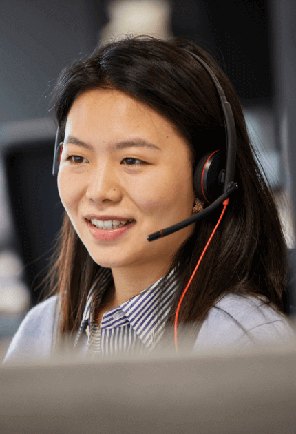 Image shows a close up of someone with long dark hair, smiling and speaking into a headset in front of a computer screen