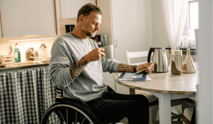 Gentleman sat at his kitchen table in his wheelchair as he enjoys a cup of tea and reads something