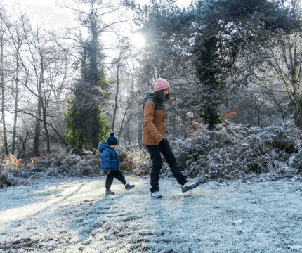 Mum stomping through frosty grass with a coat and hat on and her young son copying her by walking behind
