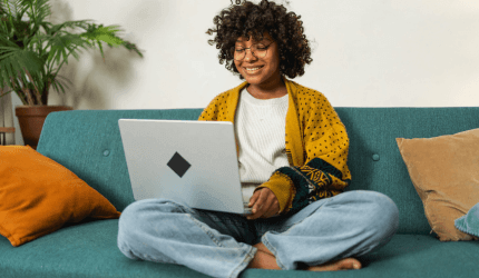An individual with short curly hair smiles whilst they use their laptop on the sofa