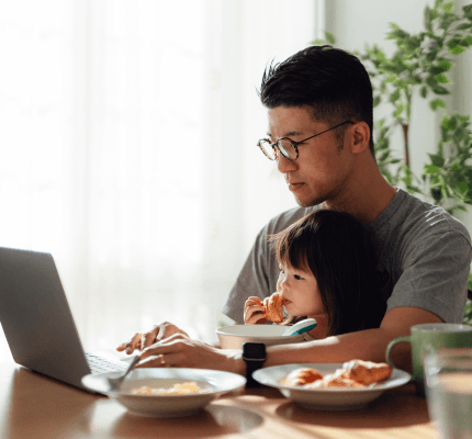A parent uses their laptop at the table whilst their young child sits on their lap eating breakfast