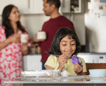 Young child smiles and paints at the table whilst their parents enjoy a cup of tea together in the background