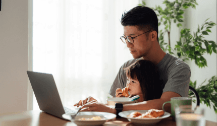 A parent sits at the table on their laptop whilst a young child sits on their lap eating breakfast