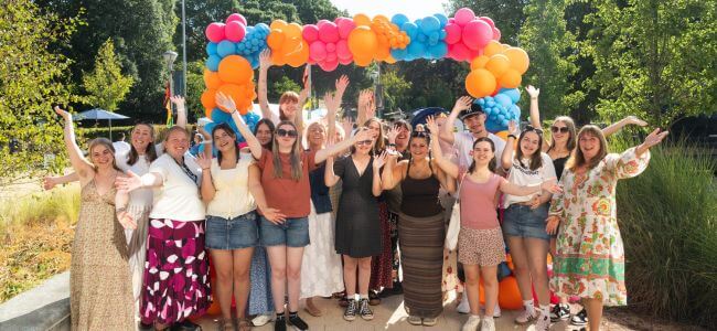Group of students smiling and waving to camera with brightly-coloured balloon arch behind