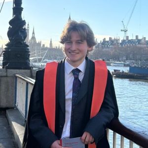 Student in graduation robes leans on rail with Thames waterside in the background