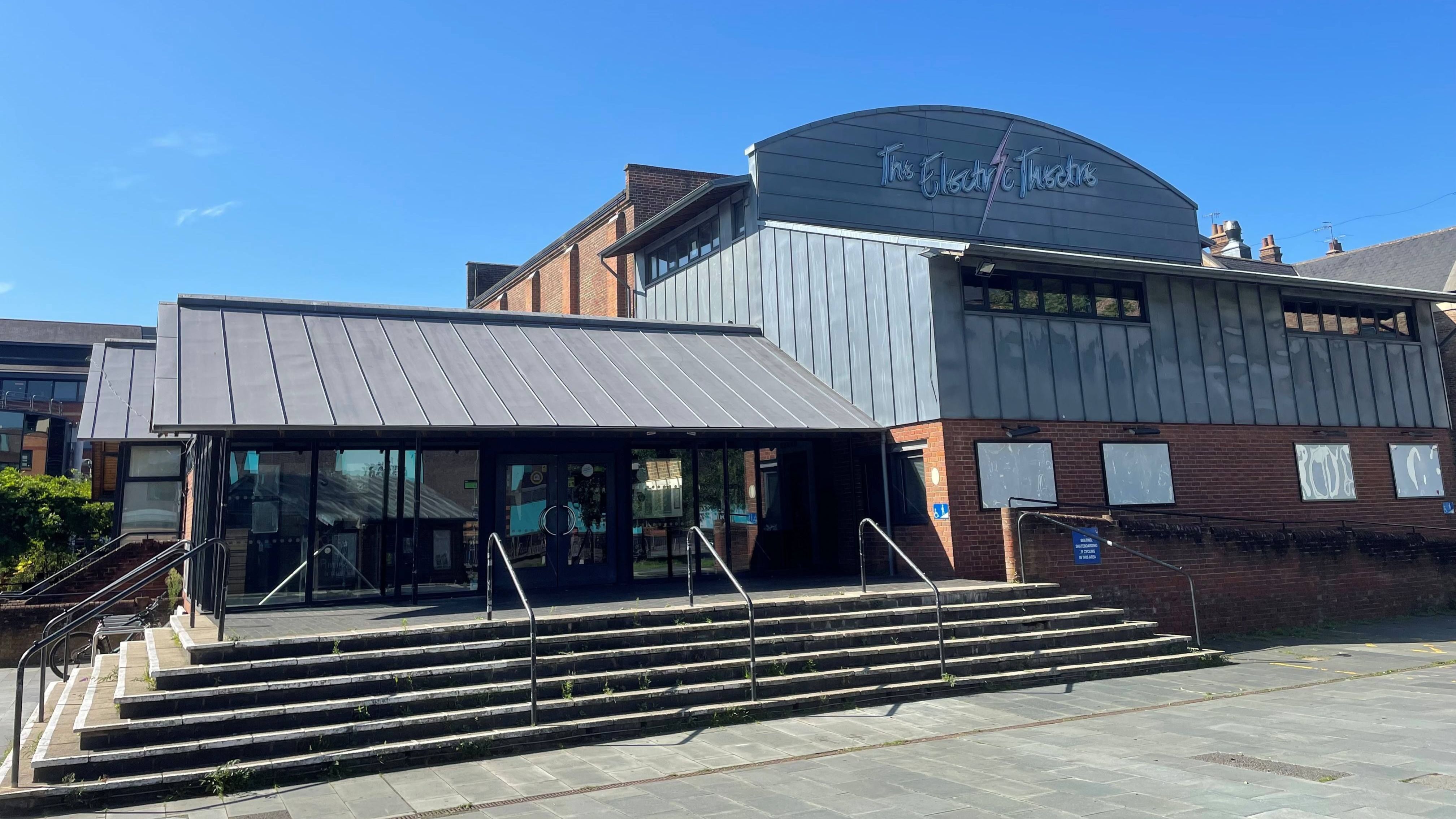 A photo of The Electric Theatre, a modern brick and metal‑clad building with a stepped entrance and handrails leading to glass double doors. The sky is bright and clear, and the theatre’s name is displayed on a curved sign near the roof