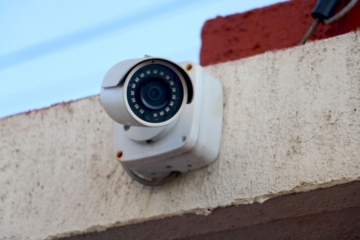 A white security camera mounted on an exterior wall, with its lens visible.