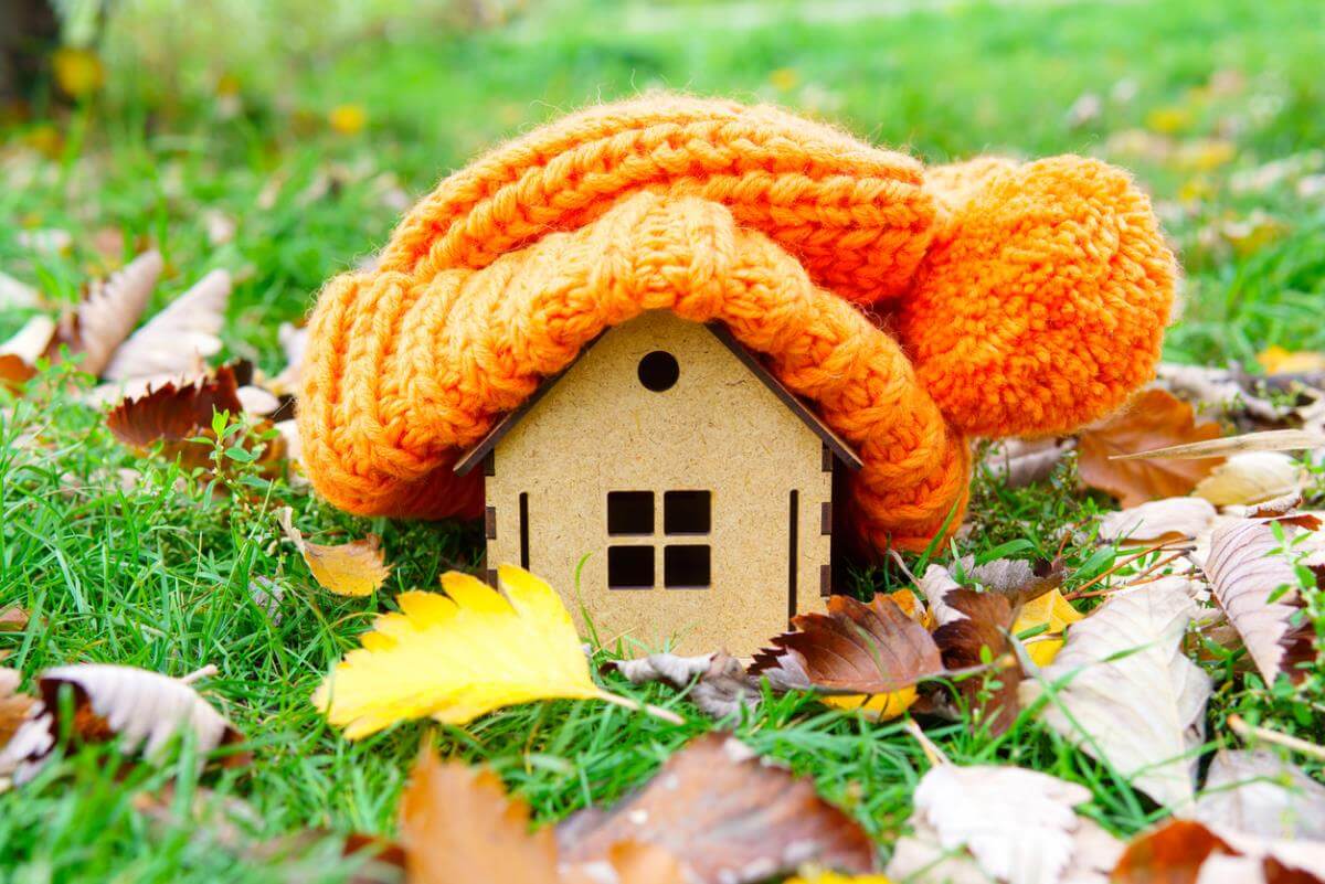 Small model house covered by a knitted orange hat on grass surrounded by autumn leaves.