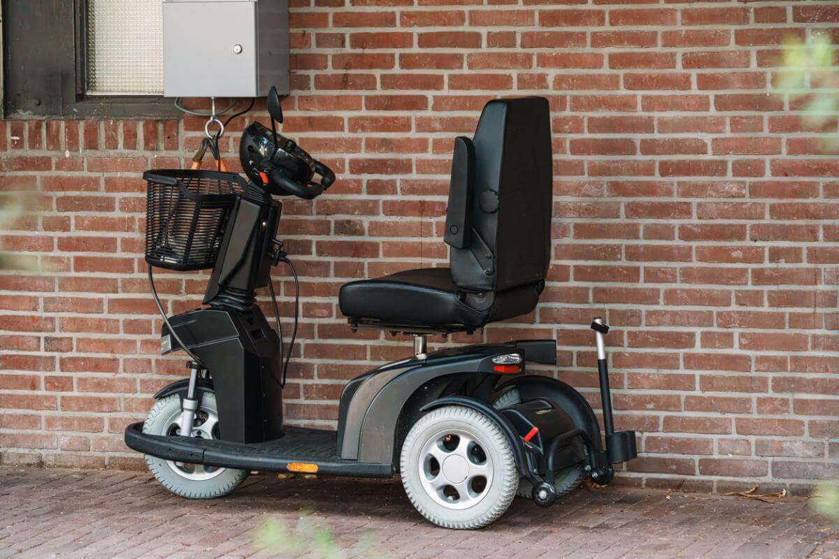 A black mobility scooter with a front basket parked on a brick-paved surface against a red brick wall.