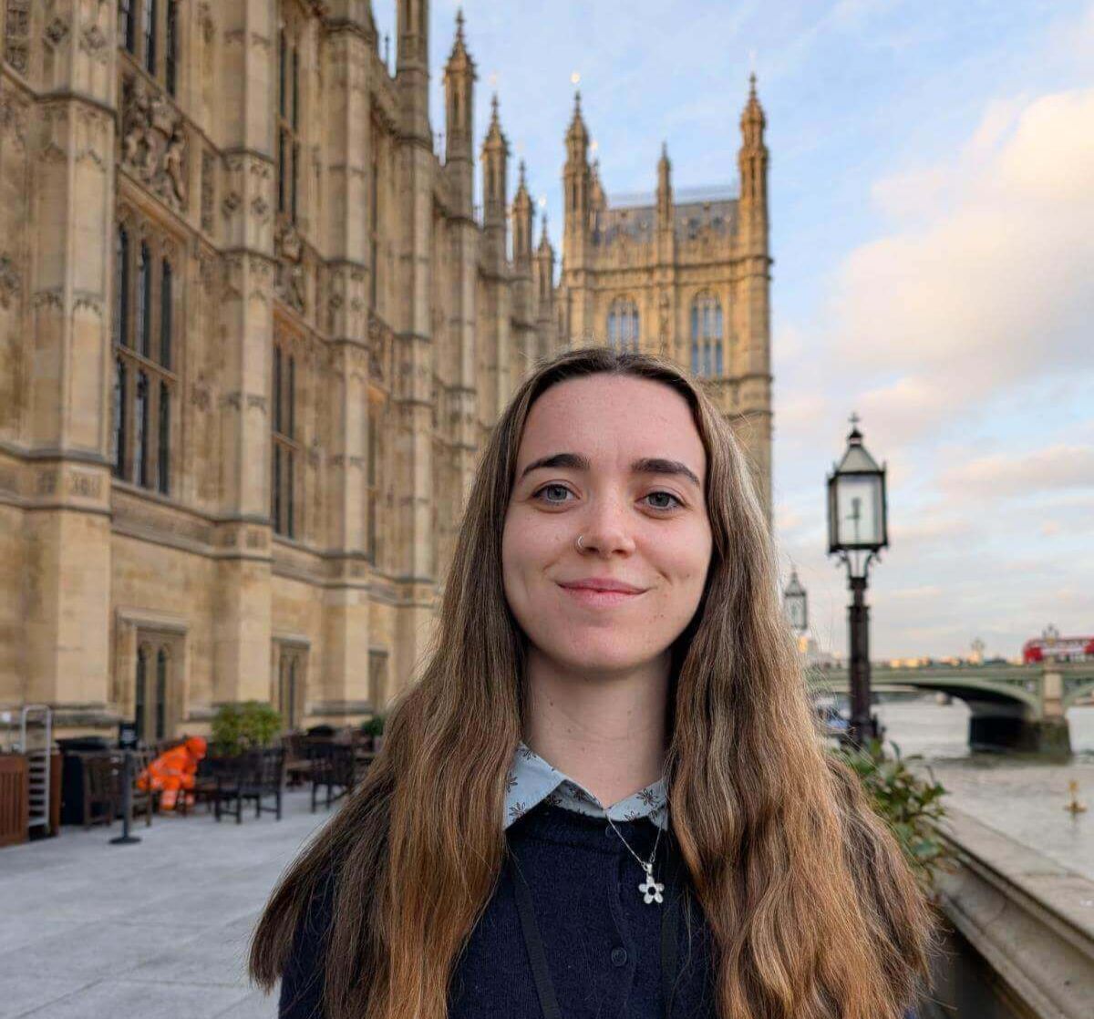 Lucy Unwin stands smiling outdoors beside the River Thames with the Houses of Parliament in the background.
