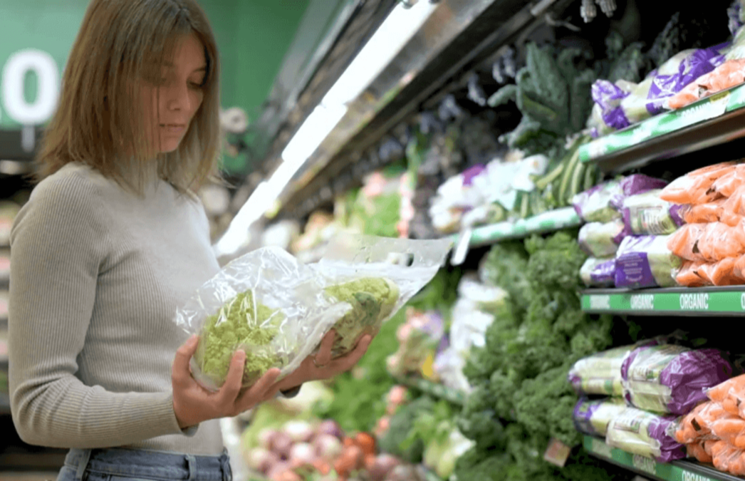 A shopper in a supermarket produce aisle examines a bag of broccoli, with shelves of leafy greens and packaged vegetables (including carrots) in the background.