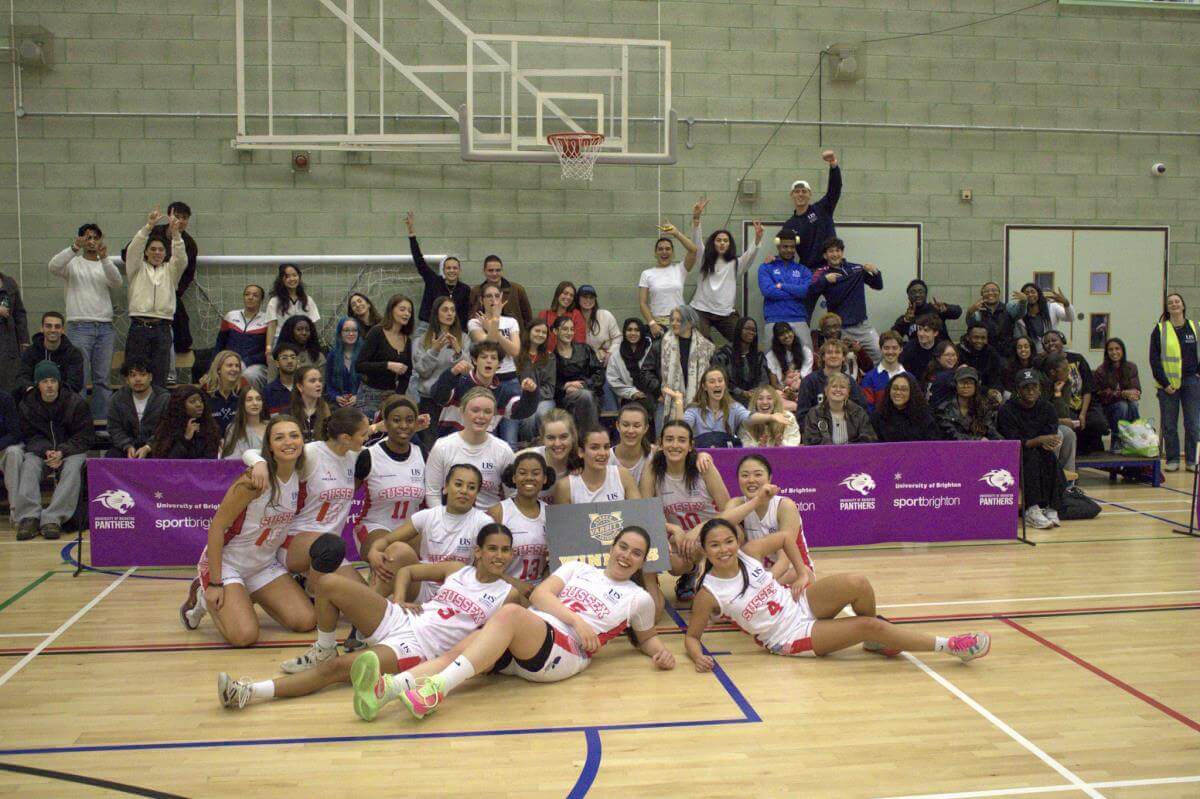 Sussex Women's Basketball team post with winners plaque and crowd cheer in background.