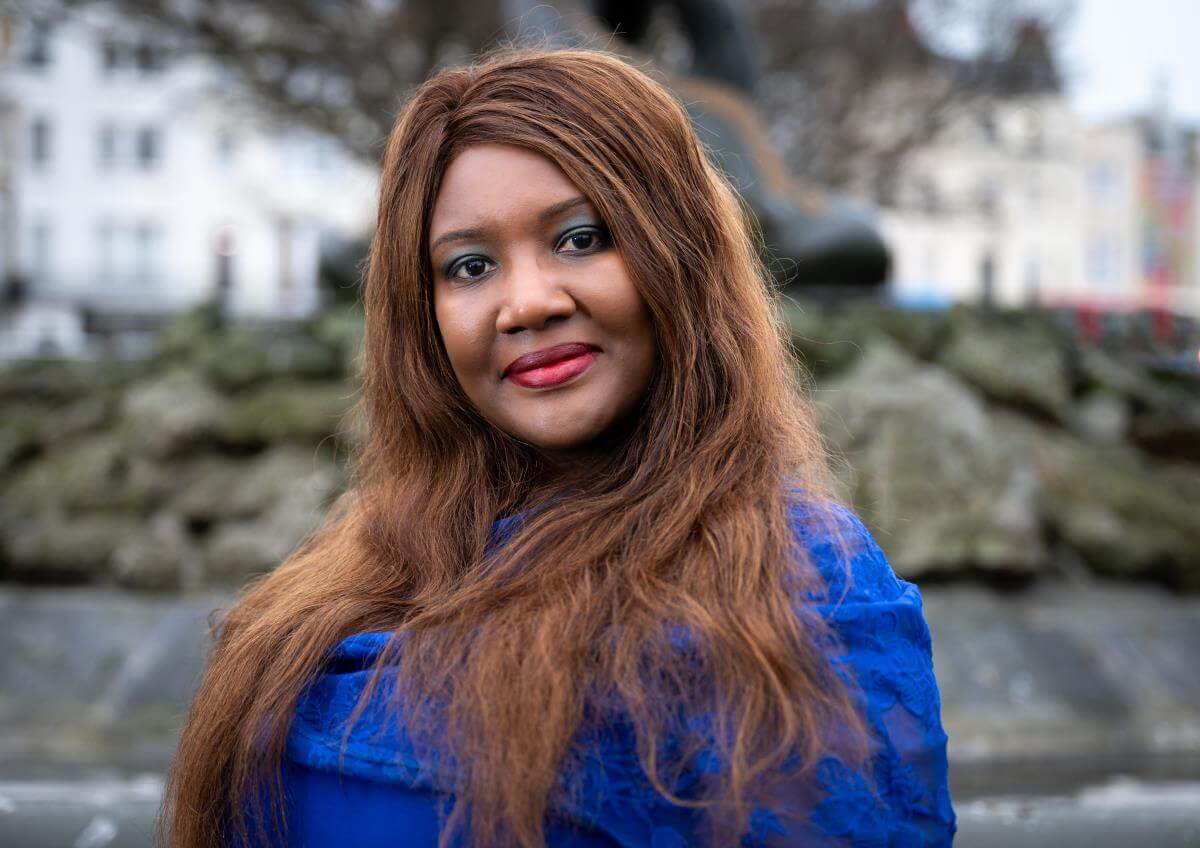 Dr Tam Cane outdoors, facing the camera and smiling, with long auburn hair and a bright blue top, set against a softly blurred street. 