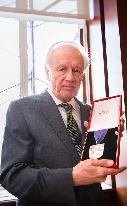 Professor Sir Anthony Leggett standing indoors by a window, wearing a suit and holding an open presentation case displaying his Nobel Prize medal.