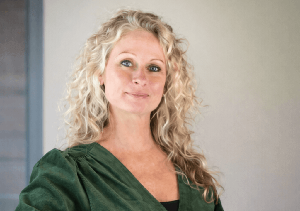Associate Professor Rebecca Stephens standing indoors, facing the camera and smiling, wearing a dark green blouse against a softly blurred neutral background.