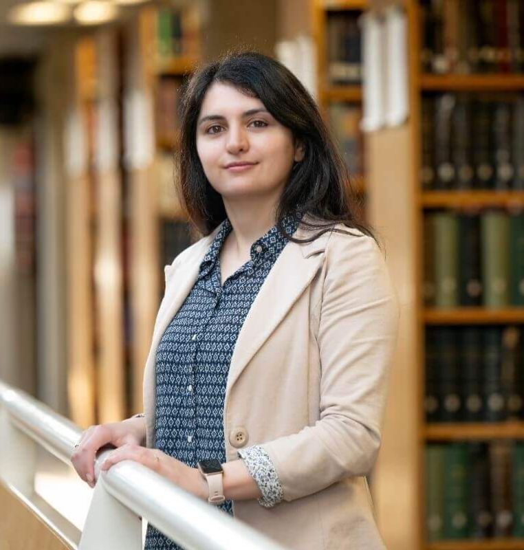 Dr Marianna standing in a library, facing the camera and smiling, wearing a beige blazer and patterned blue blouse, with blurred bookshelves in the background.