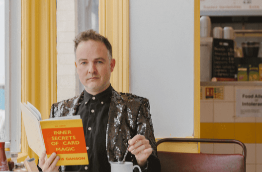 Dr Augusto Corrieri (Vincent Gambini) seated in a café, looking at the camera while holding a bright orange book titled Inner Secrets of Card Magic.