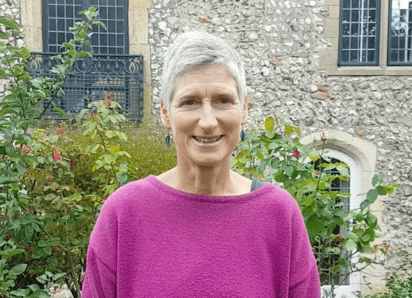 Professor Gail Davey standing outdoors and smiling at the camera, wearing a bright pink jumper, with a stone building and leafy garden in the background.