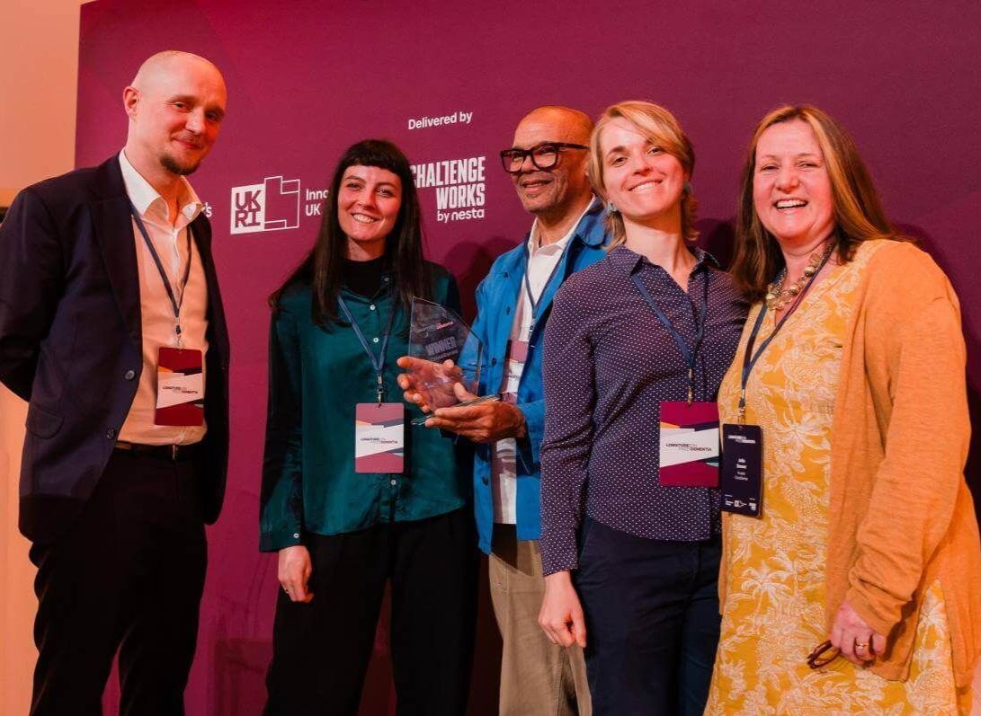 Professor Julia Simner, far right, and the Animorph team stand smiling on stage at the Longitude Prize on Dementia awards, with one team member holding the winner’s trophy in front of a purple event backdrop.