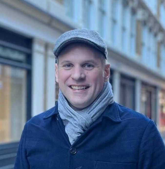 Dr Felix Roesch smiling at the camera on a city street, wearing a grey flat cap, grey scarf and dark blue coat, with buildings softly blurred in the background.