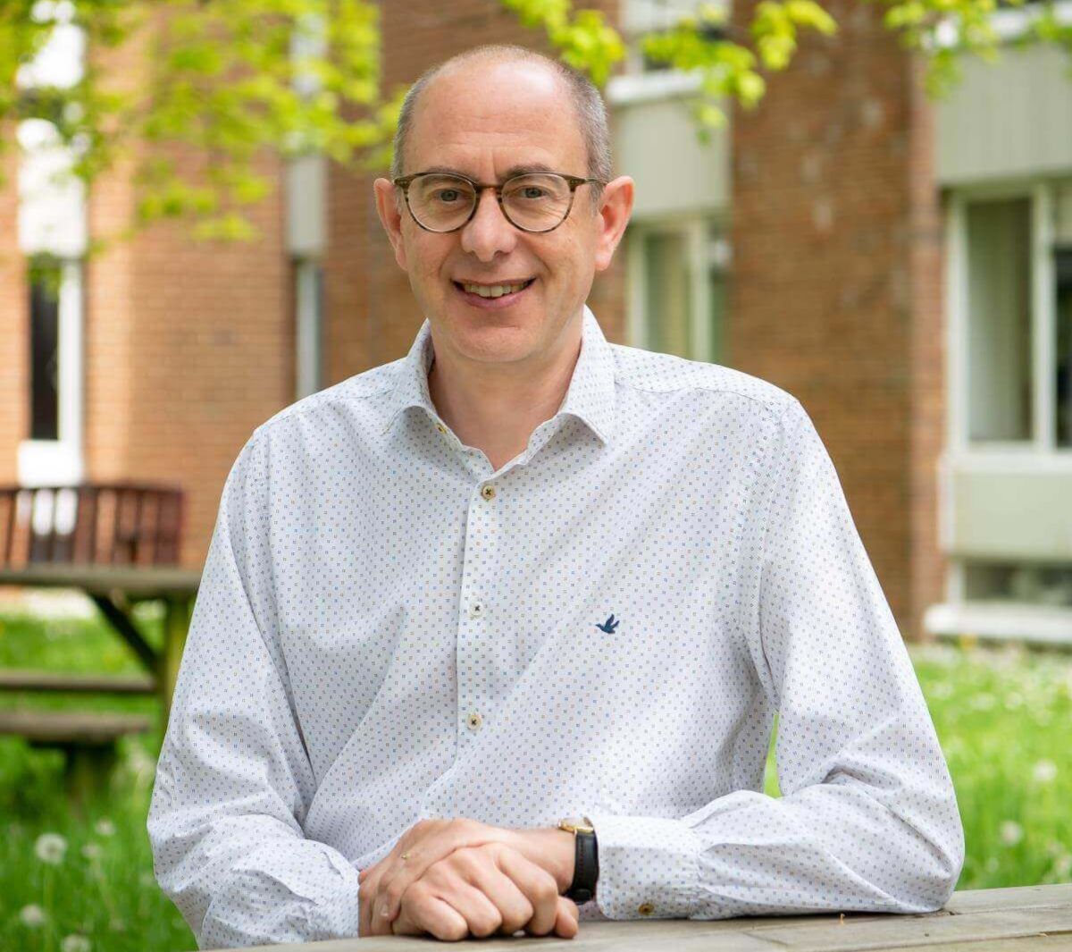 Professor Geert De Neve smiling at the camera outdoors, wearing glasses and a white patterned shirt, seated at a wooden table with campus buildings and green trees softly blurred in the background.