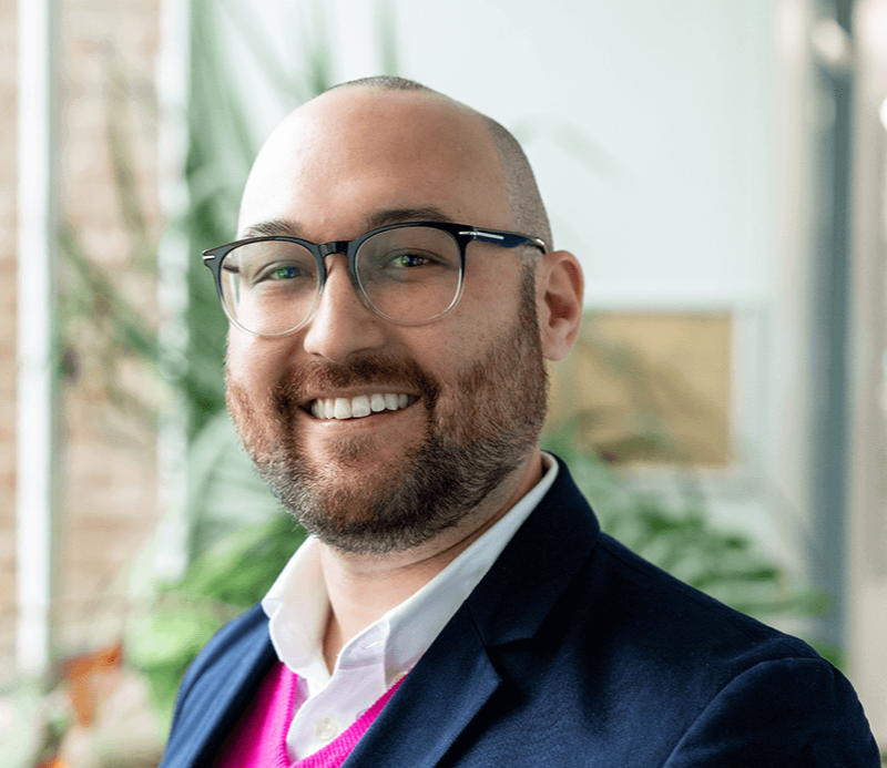 Dr Luke Yates smiling in an indoor portrait, wearing glasses, a navy jacket, white shirt and bright pink jumper, with plants and softly blurred windows in the background.