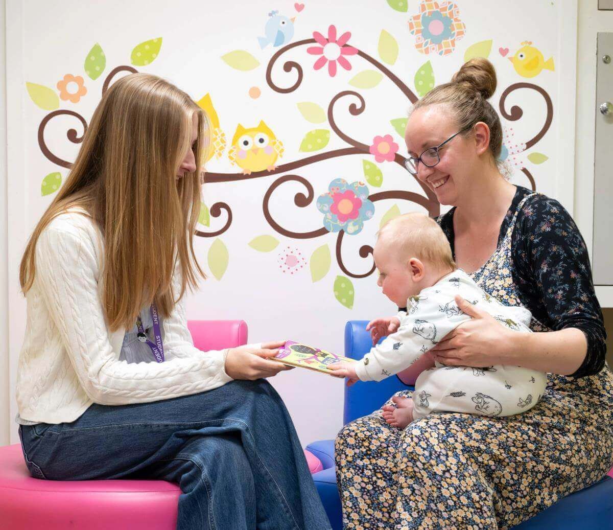A researcher shows a brightly coloured board book to a baby sitting on an adult’s lap in a child-friendly room, with pink and blue seats and a wall mural of flowers, leaves and owls in the background.