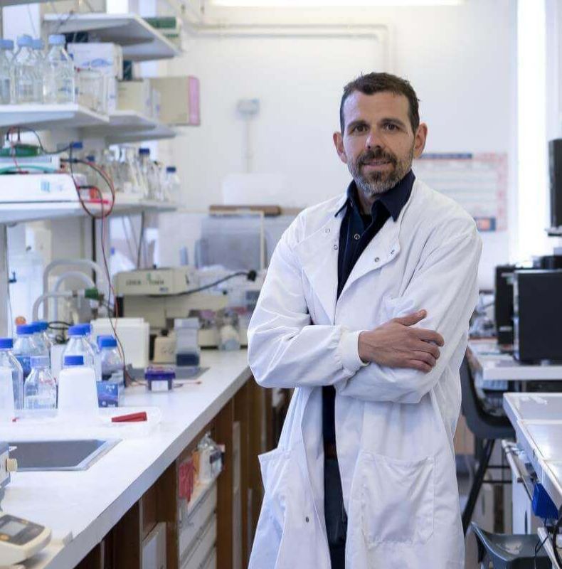 Professor Georgios Giamas stands in a laboratory wearing a white lab coat, surrounded by lab benches, bottles and scientific equipment.