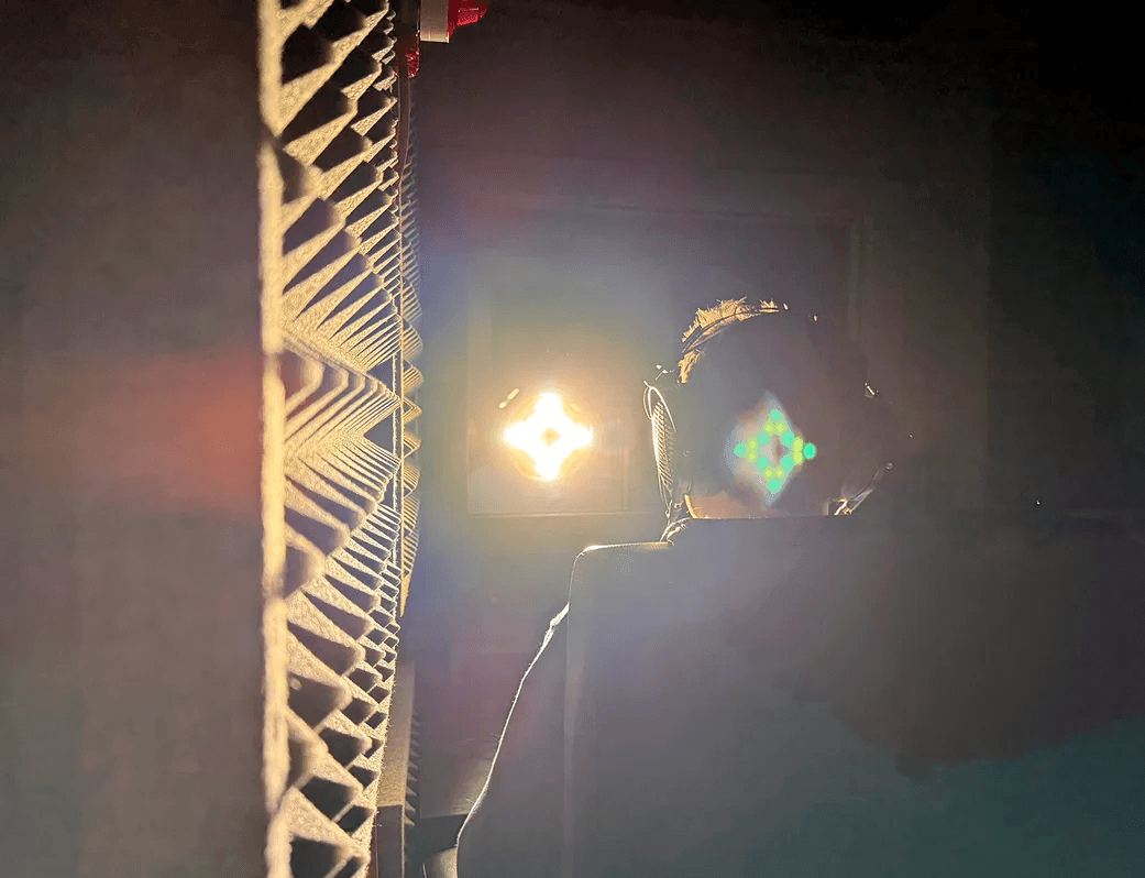 A person sits in a dark soundproof booth wearing headphones, with a bright strobe light shining towards their closed eyes. A foam acoustic panel is visible on the left.