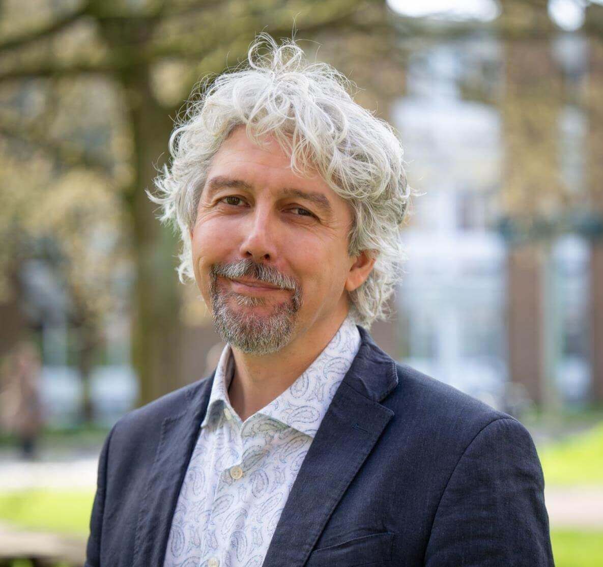 Professor Michael Collyer smiling outdoors, wearing a navy blazer and light shirt, with trees and campus buildings softly blurred in the background.
