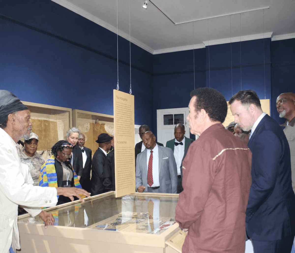 Scobie Lekhutile of Khama III Memorial Museum speaks to guests gathered around a museum display case showing repatriated cultural belongings at the official exhibition launch in Botswana. Professor JoAnn McGregor is pictured fourth from left.