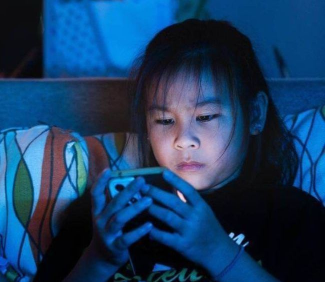 A young child lying on a sofa in a dark room, looking at a smartphone illuminated by blue screen light.