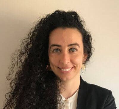 Portrait of Dr Özden Melis Uluğ smiling at the camera, wearing a dark blazer and white shirt against a plain light background.