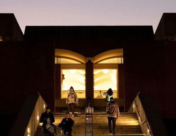 People walk up the steps outside the University of Sussex Library at dusk, with the warmly lit entrance windows shaped like an open book.