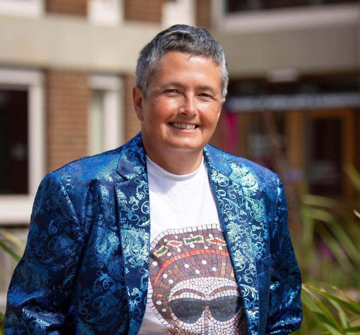 Professor Liz James smiles outside on campus, wearing a bright blue patterned jacket over a T-shirt featuring a mosaic-style design.